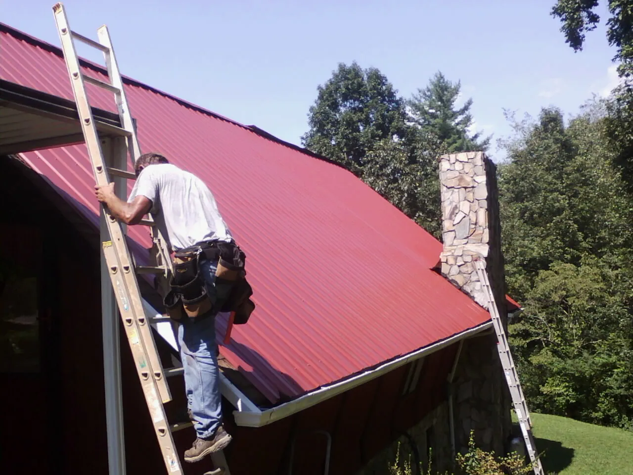 Roofer on a ladder beside a red metal roof and stone chimney — Soffit & Fascia Repair by Roofing Brothers in Stratham
